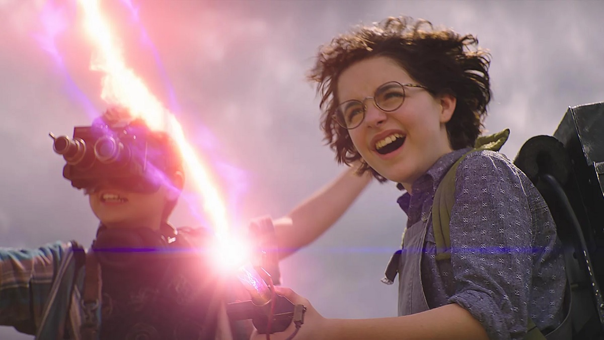 Phoebe (Mckenna Grace) grins with delight as she fires a proton pack, while Podcast (Logan Kim), wearing a pair of night vision goggles, cheers her on in Ghostbusters: Afterlife.