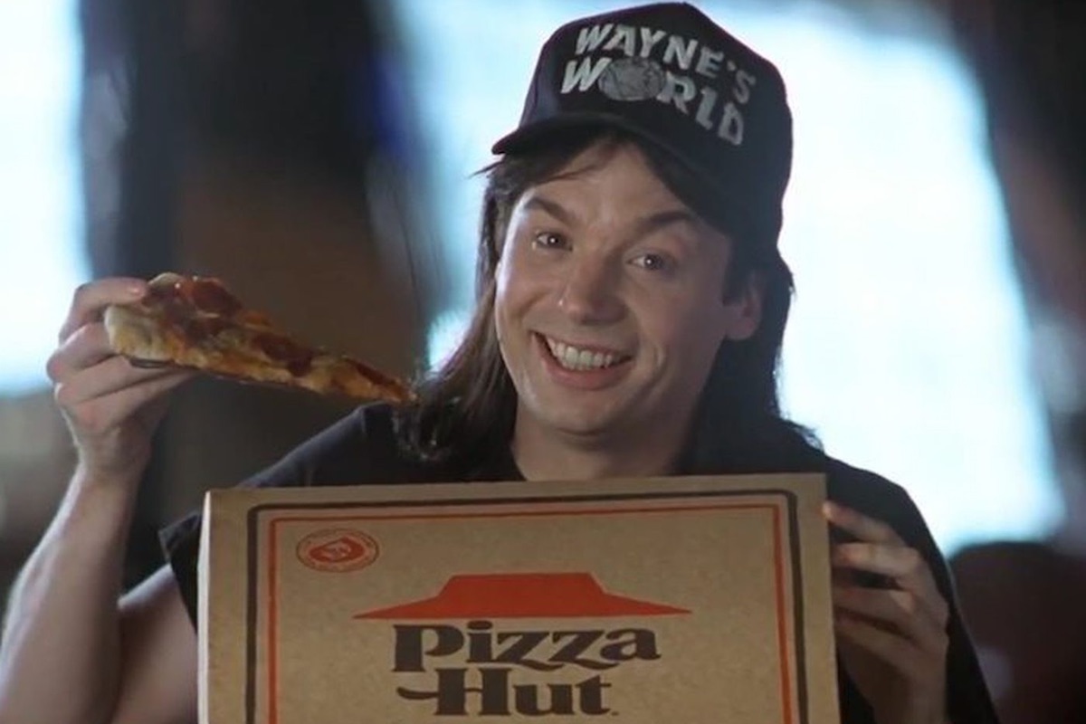 A still from Wayne's World shows Mike Myers as Wayne eating a slice of Pizza Hut pizza