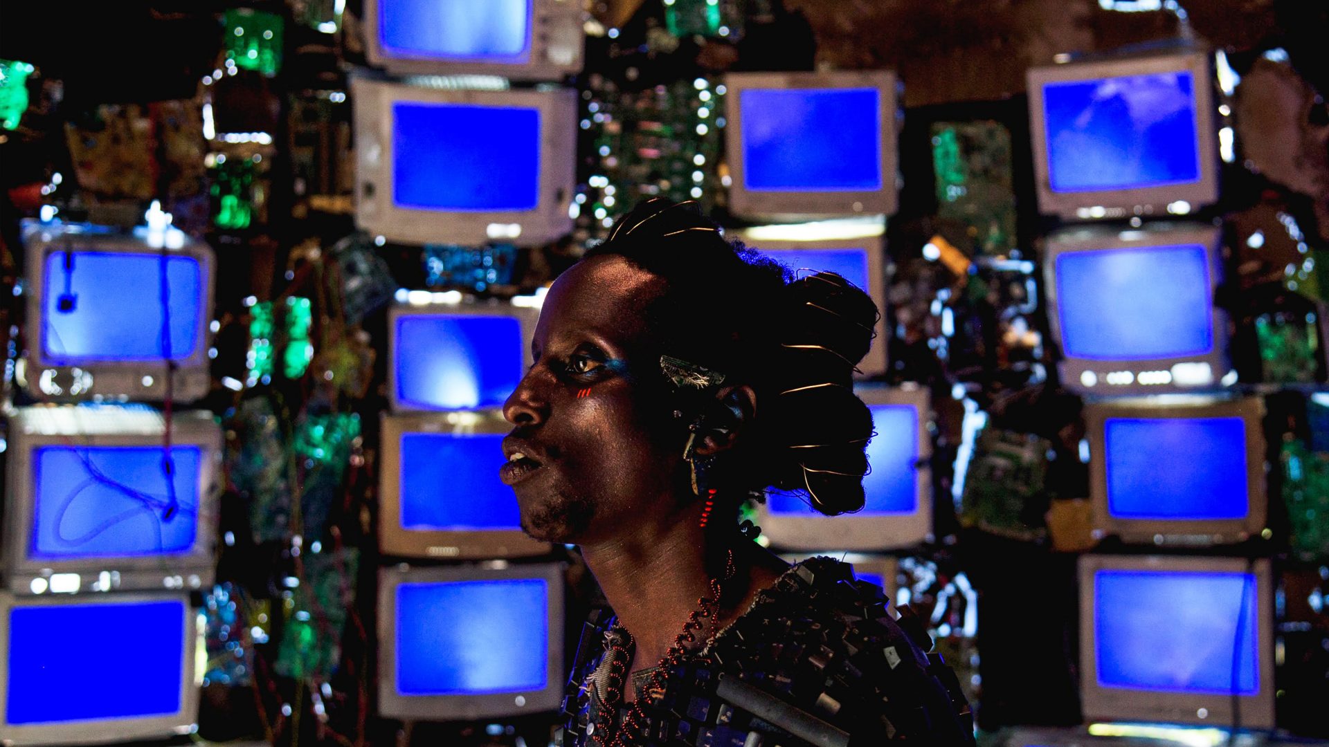 a still of a Rwandan person wearing Afrofuturist clothing in front of TV screens in Neptune Frost