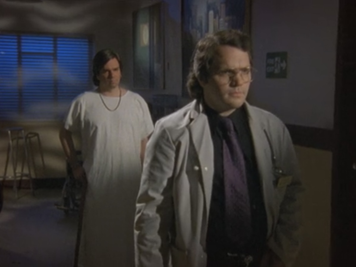 Sanch in a robe and a cane stands behind Rick Dagless on Garth Marenghi's Darkplace 