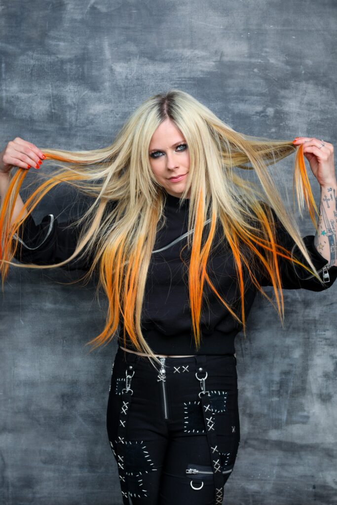 Avril Lavigne, wearing all black, plays with her blond hair with orange highlights.