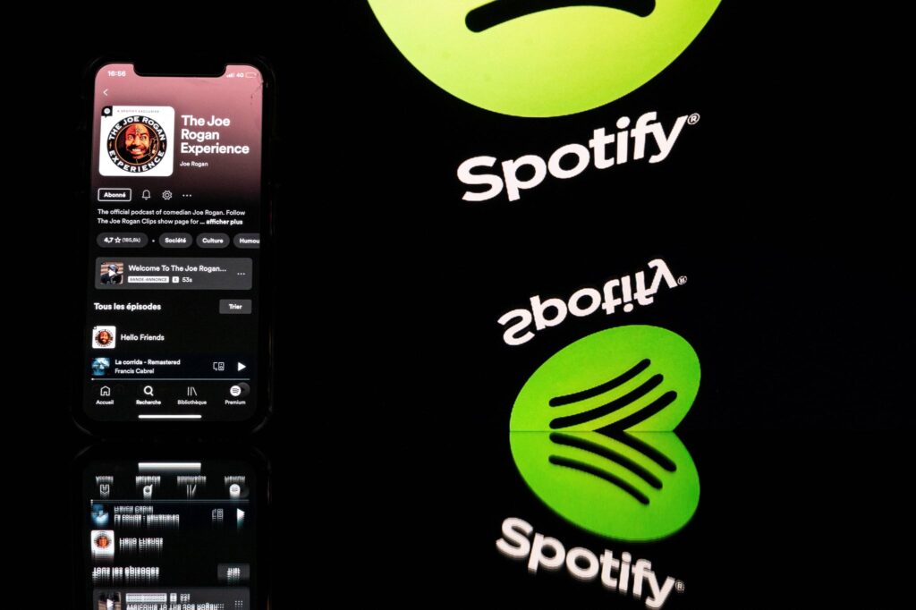 This picture taken in Toulouse, southwestern France, on January 31, 2022 shows a smartphone displaying the Joe Rogan podcast and a screen displaying the Spotify's logo. - Caught in a disinformation maelstrom, Spotify announces it will guide listeners of Covid-19 podcasts to established facts about the pandemic. This comes after artists pulled their songs in protest at the platform given on Spotify to podcaster Joe Rogan, accused of spreading damaging myths about the pandemic. (Photo by Lionel BONAVENTURE / AFP) (Photo by LIONEL BONAVENTURE/AFP via Getty Images)