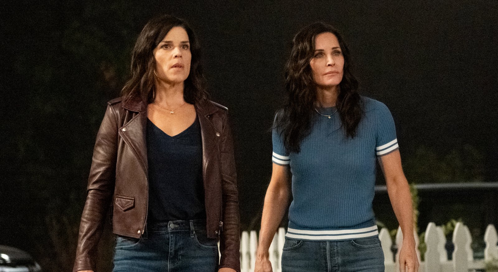A still from Scream (2022) shows Neve Campbell and Courtney Cox as Sydney Prescott and Gale Weathers standing in front of a white picket fence