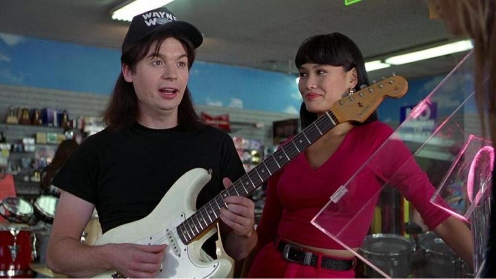 A still from Wayne's World shows Mike Myers as Wayne holding a white electric guitar standing next to Tia Carrere as Cassandra in a guitar shop