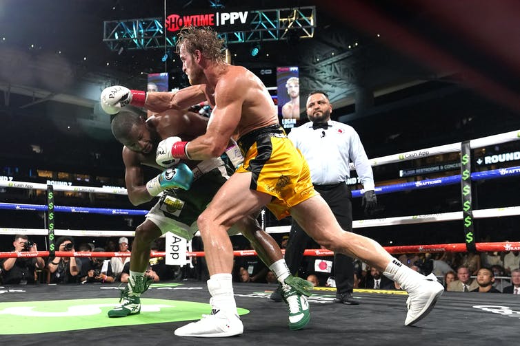 Two men box in a boxing ring with a referee in the background, the man in the foreground throws a right hook towards his ducking opponent