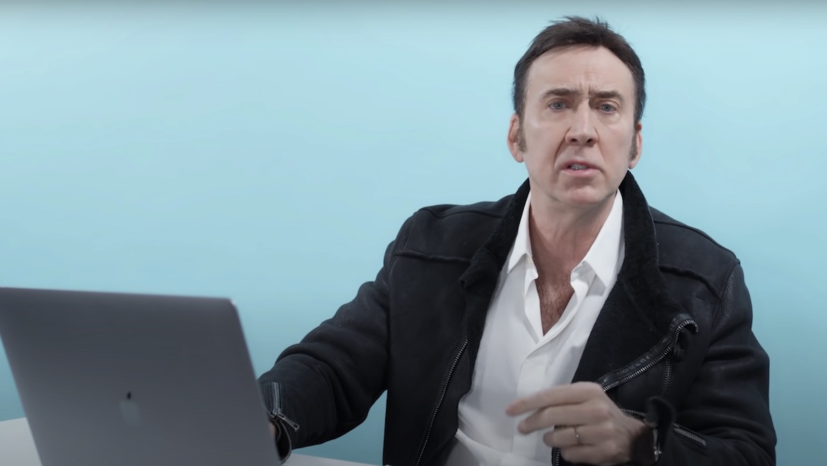 Nicolas Cage sin a white shirt and black jacket sitting at a laptop