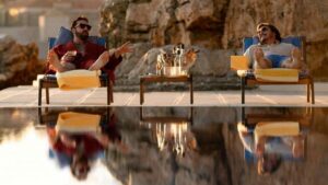 A still from The Unbearable Weight of Massive Talent shows Nic Cage as Nick Cage and Pedro Pascal as Javi laying on pool loungers in Mallorca, Spain