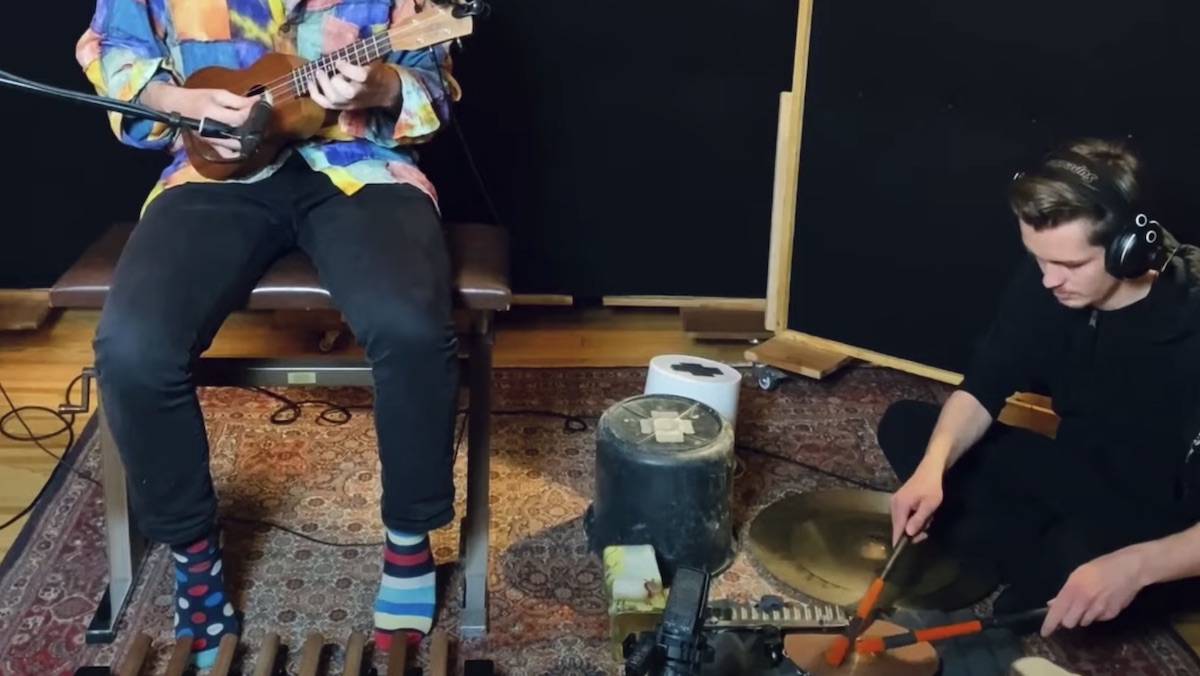 A man sits on the floor of a recording studio playing drums while another (face unseen) sits on a stool playing a ukulele