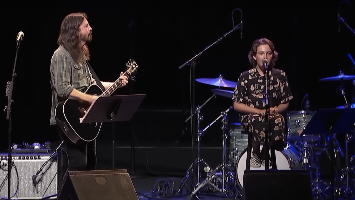 Dave Grohl plays a guitar on stage while his daughter Violet sits behind a mic singing