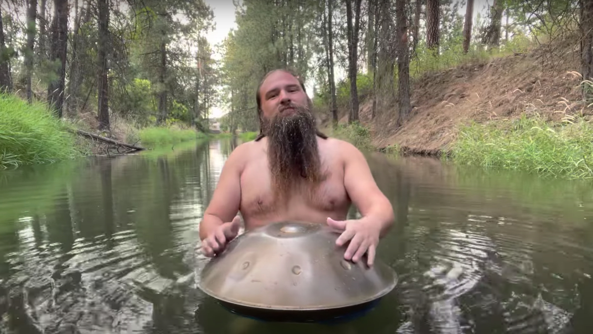 A man with long hair and no shirt on playing a handpan in a river in the woods.