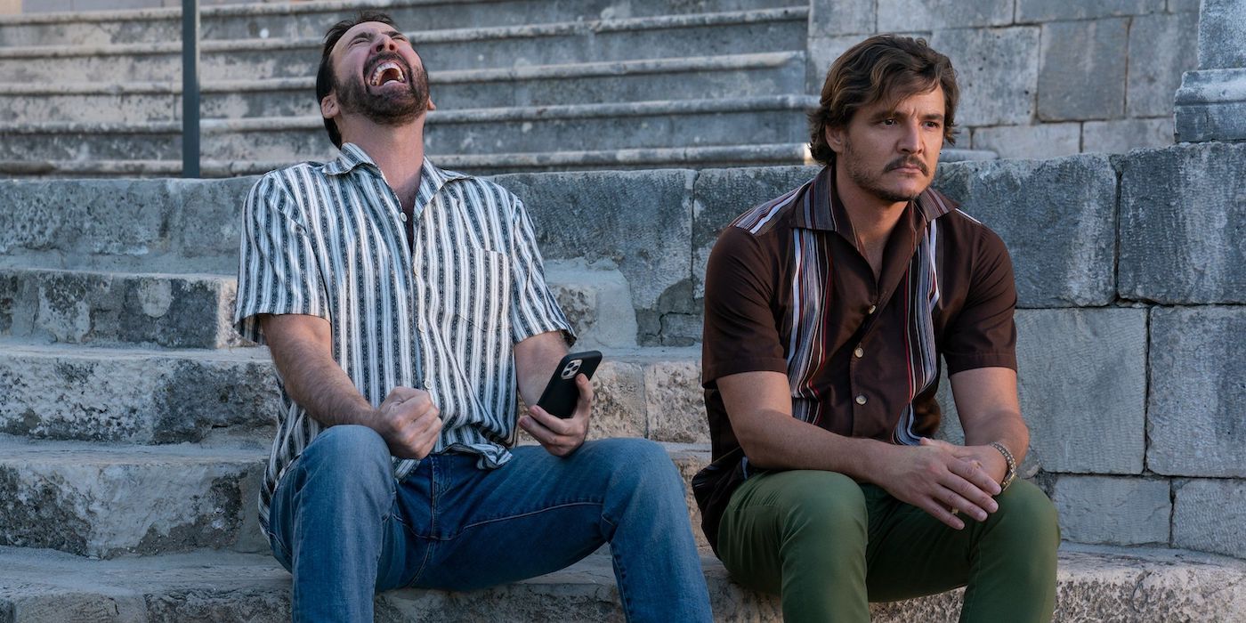 A still from The Unbearable Weight of Massive Talent shows Nic Cage as Nick Cage and Pedro Pascal as Javi sitting on a set of stairs as Cage laughs maniacally 