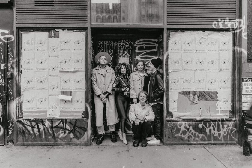 Five members of a rock band stand in front of a graffitied doorway