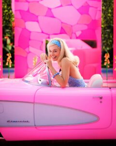 Margot Robbie as Barbie the doll from the titular film, sitting in a miniature Cadillac, smiling.