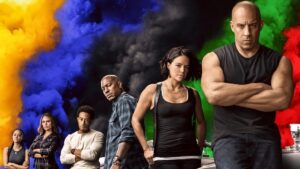 The Fast and the Furious cast stand in a diagonal line against a row of cars while different color smoke goes up behind them. Fast X has a new director in Louis Leterrier.