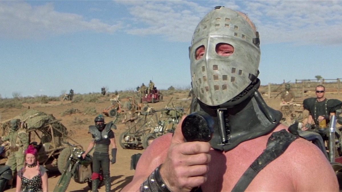 The Lord Humongous with a hockey mask and leather straps, speaks into a microphone in The Road Warrior.