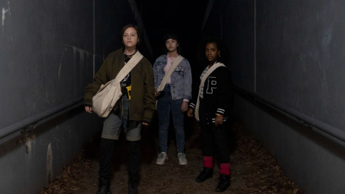 tiffany, mac, and erin stand in a dark area wearing paper girls bags tv show