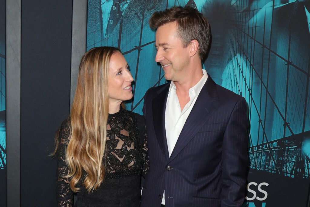 Shauna Robertson (L) is wearing a black, lace-patterned dress and smiling up at Edward Norton, who is looking back at her. He is wearing a dark blue blazer over a white dress shirt.