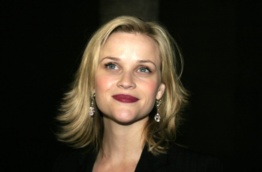 Closeup of Reese Witherspoon against black background in black top. She is wearing dangle earrings and deep red lipstick