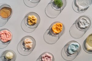 Multicolored creams and scrubs in many Petri dishes on gray background. Concept of cosmetics laboratory researches. Photography in flat lay style