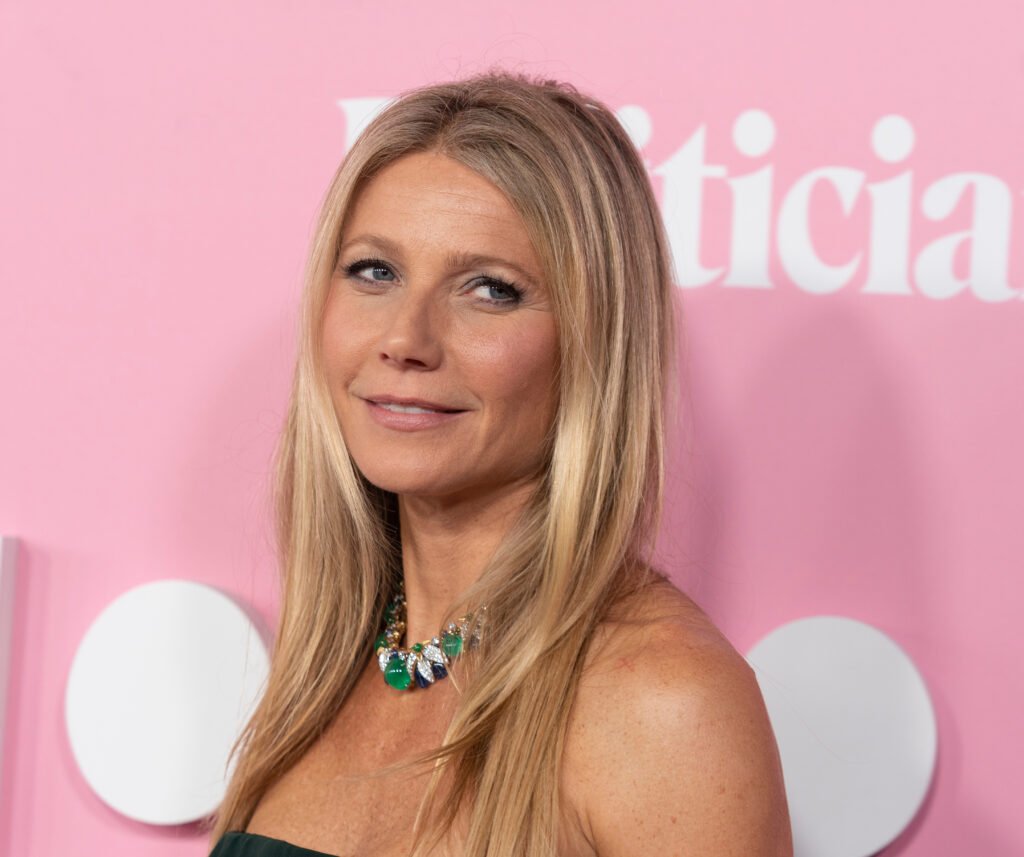 Gwyneth Paltrow smiles in strapless black gown with green necklace against bubblegum pink backdrop