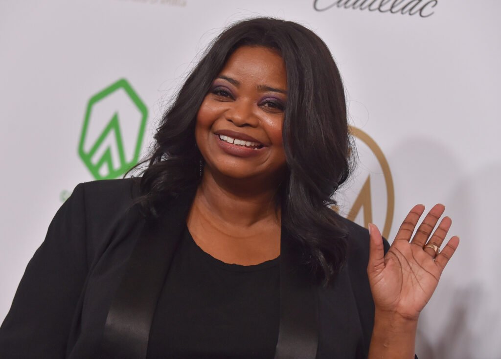 Octavia Spencer waving and smiling in black top and black blazer against white backdrop