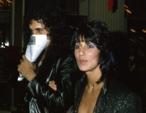 Gene Simmons (L) holding piece of paper over his face and wearing leather jacket, walking with Cher, who is wearing a sparkly black outfit with bold purple eye makeup