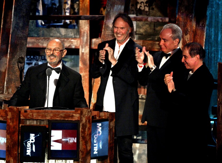 A man in a tuxedo stands in front of a podium, as three other men stand nearby and applaud.