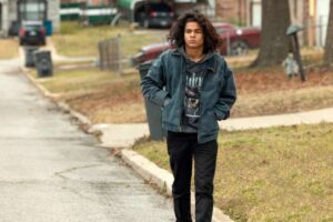 A long-haired teen boy in a denim jacket walks down a street on a gray day.
