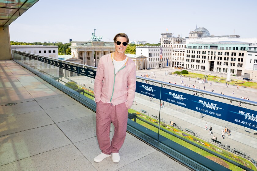 A man in a pink cardigan with hands in pockets of rose pants stands in front of a glass railing