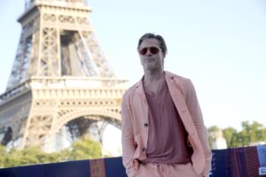 A man in a pink suit and rose shirt poses in sunglasses in front of the Eiffel Tower