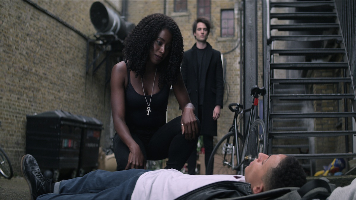Death kneels over a dead man in an alley as Morpheus looks on in The Sandman