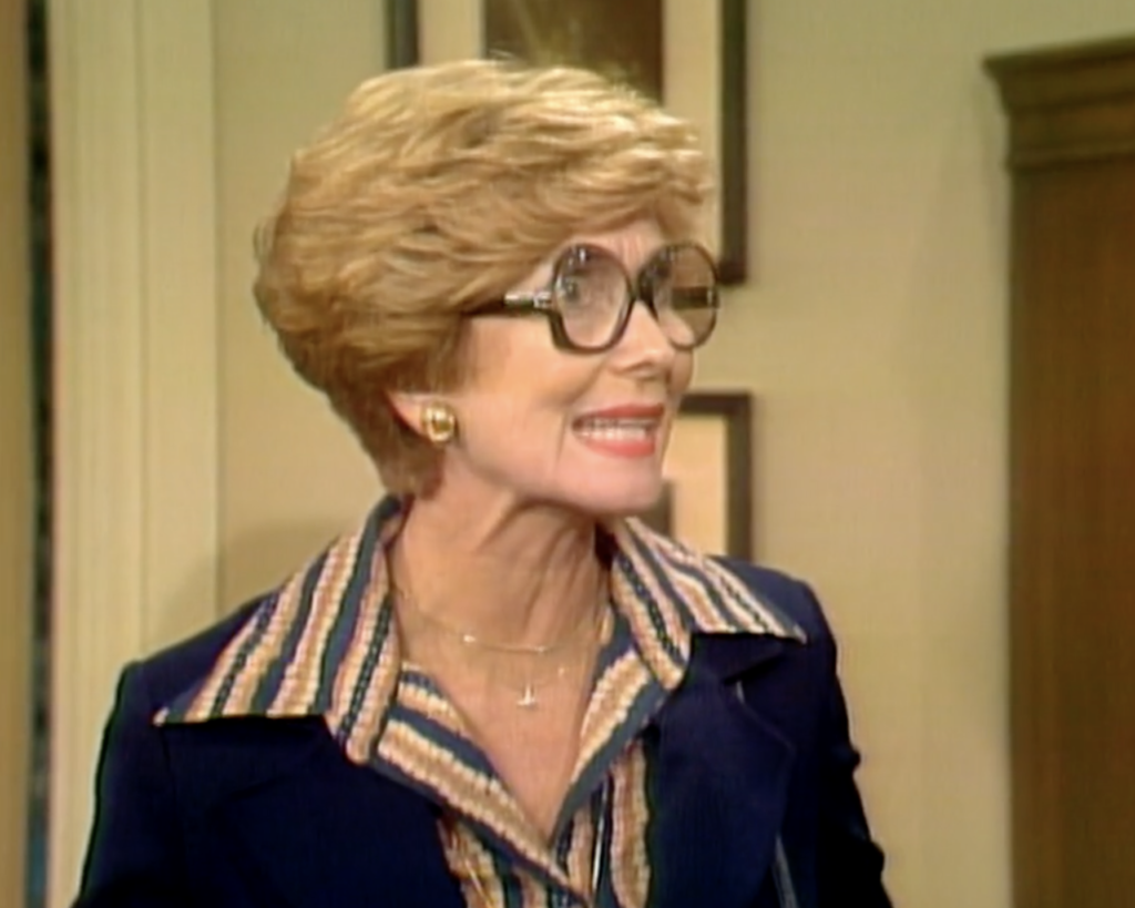 Actress Sheila Rogers in "Three's Company," wearing big round glasses and a striped dress shirt