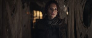 Jyn Erso, a dark haired woman wearing a jacket and scarf, emerges from the shadows in Rogue One: A Star Wars Story