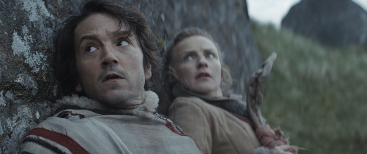 A close-up on Cassian and Vel hiding behind a rock, and looking shocked at something out-of-frame