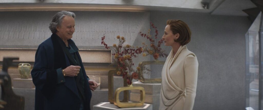Luthen talking to Mon Mothma in a shop, with him standing in front of her. Flowers are in vases on the table framed between them in the background