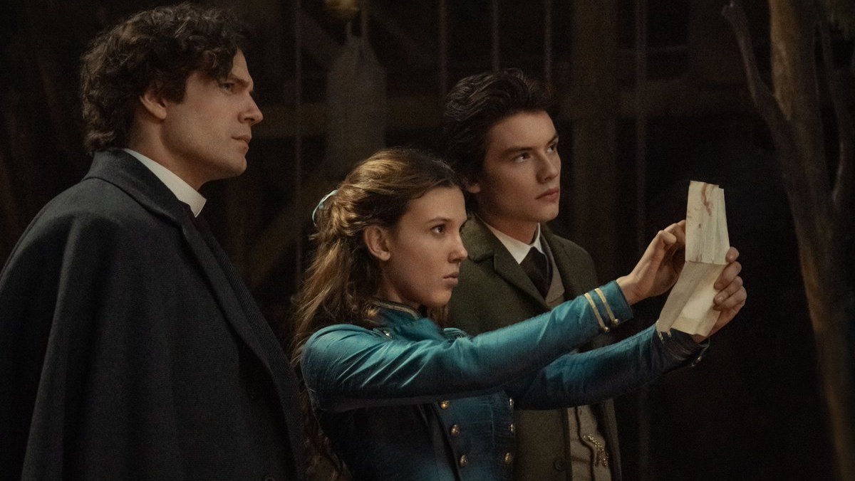 Enola Holmes (Millie Bobby Brown) holds up a piece of evidence with Sherlock (Henry Cavill) to her right and Tewkesbury (Louis Partridge) to her left.