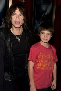 Steven Tyler (L) in black leather jacket standing next to his young son, who is wearing a red tee shirt