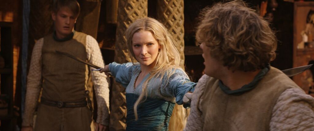 Galadriel, in a blue dress, stands between two opponents pointing a sword at both of them with something approaching a grin on her face.