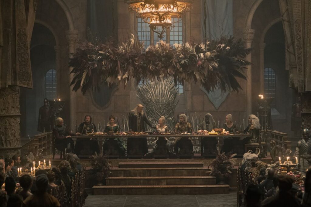A still of the royal table in the great hall of King’s Landing, with (from left to right): Daemon, Ser Strong, Alicent, Viserys, Rhaenyra, Laenor, Corlys, Rhaenys, and Laena around it facing other tables below them. Viserys is standing and looking at Rhaenyra with his arm on her shoulder.