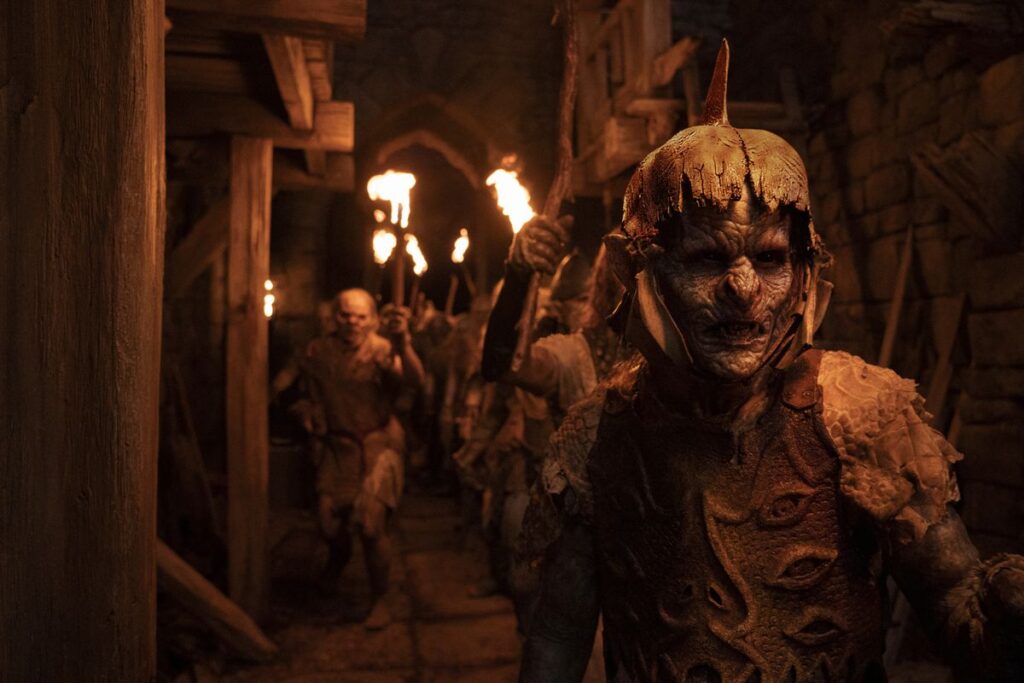 An orc soldier in the foreground of a dimly-lit, red-tinted tunnel, with orc soldiers behind him holding up torches