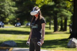 Photo of a person standing in a parking lot wearing a white virtual reality headset and holding two controllers in their hands.