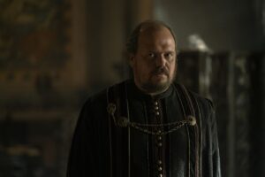 Gavin Spokes, an older man with a receding hairline, as Lord Lyonel Strong. He wears a formal Westerosi outfit and stands in a room in House of the Dragon.