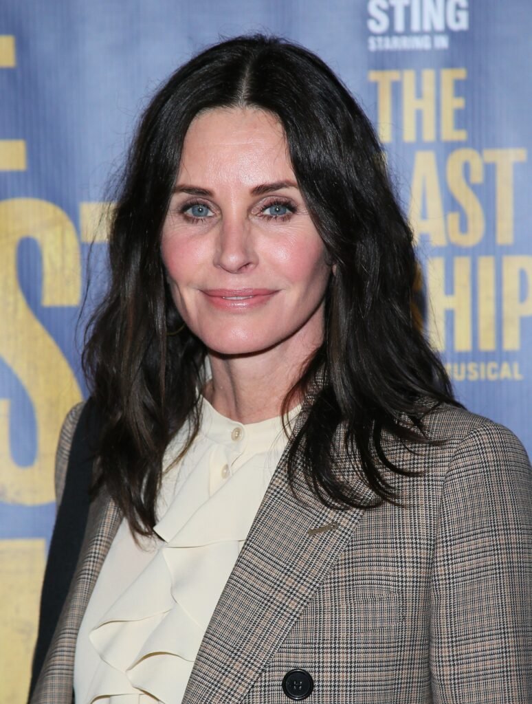 Courteney Cox smiles in cream-colored blouse with gray and tan plaid blazer