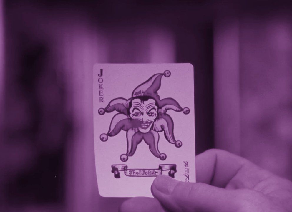 A purple tinted still of a man holding up a Joker playing card in Batman: The Silent Motion Picture.