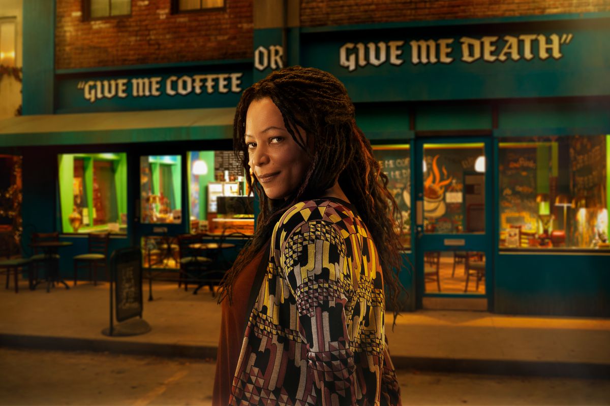 a woman with dark dreadlocks smirks in front of a shop that says GIVE ME COFFEE OR GIVE ME DEATH