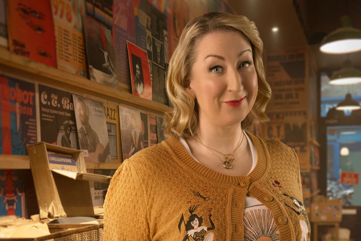 Maggie, a blonde woman in a yellow cardigan, standing in a record shop in Good Omens