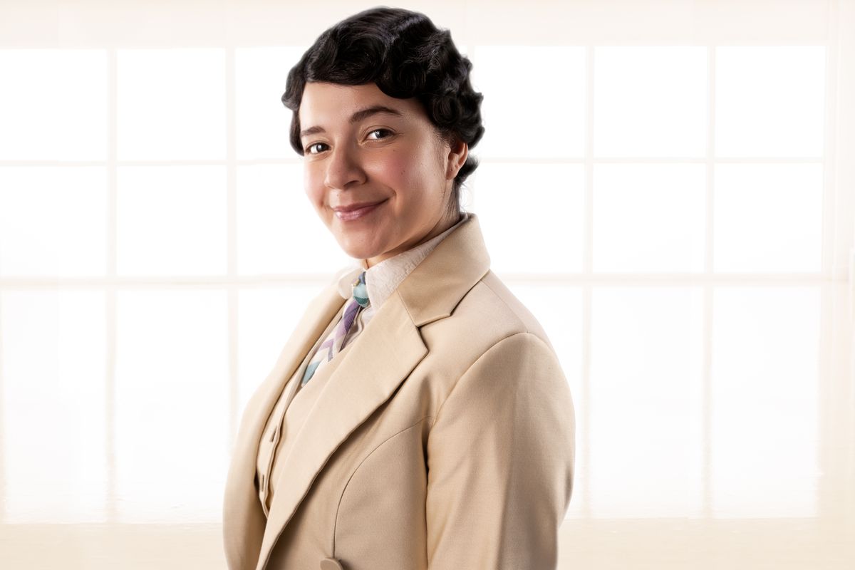Muriel, a woman with dark hair in a pale suit, standing in a cream-colored room in Good Omens
