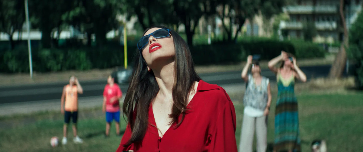 A woman in red wears dark glasses and looks at the sun, as people behind her in a park also look up, in Dark Glasses.