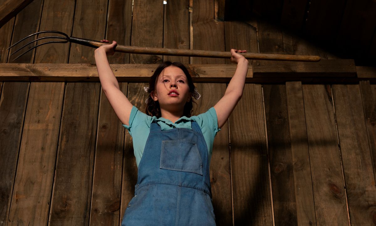 Mia Goth, wearing overalls and a blue work shirt, raises a pitchfork over her head in Ti West’s Pearl