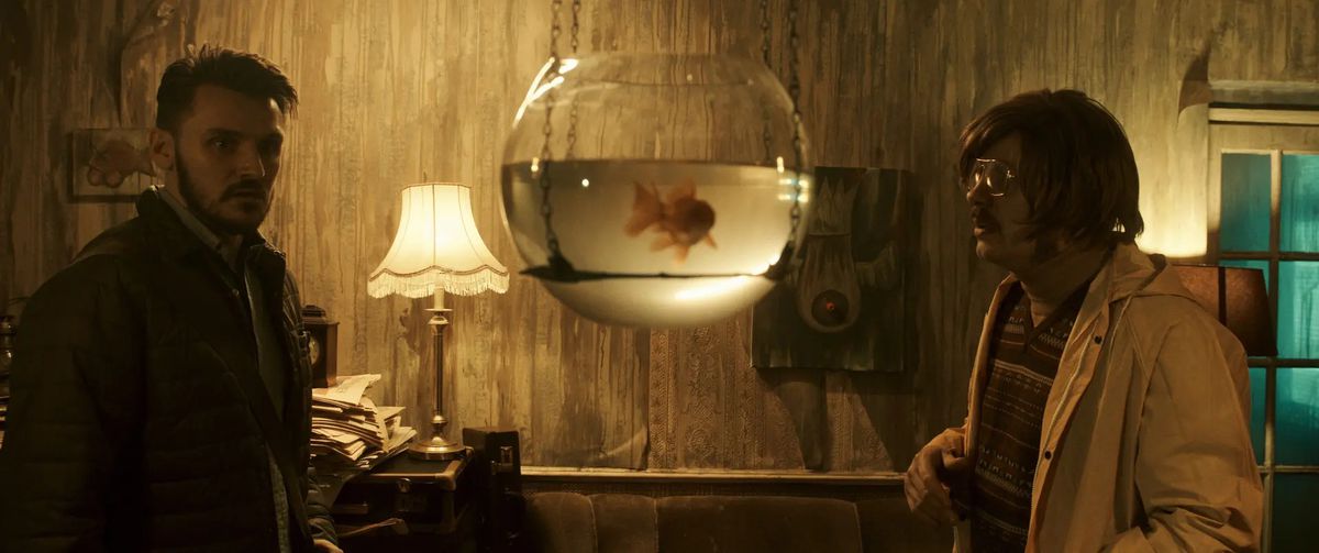 Two men stand across from one another in a dilapidated room staring at a goldfish bowl hanging from two chains in the ceiling.